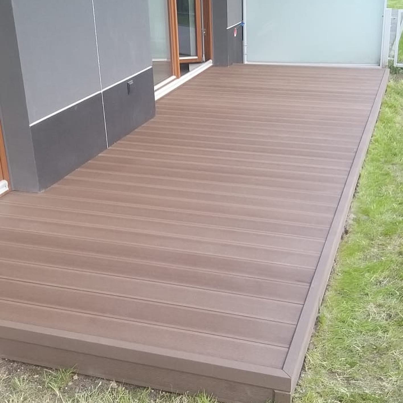 Nowoczesny brązowy taras z desek ułożonych w poprzek, przylegający do szarej ściany budynku z dużym oknem, otoczony świeżą trawą.
