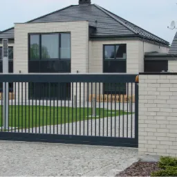 Nowoczesna, przesuwna brama w kolorze antracytowym przed domem z jasnej cegły, widok z poziomu chodnika wyłożonego kostką brukową.