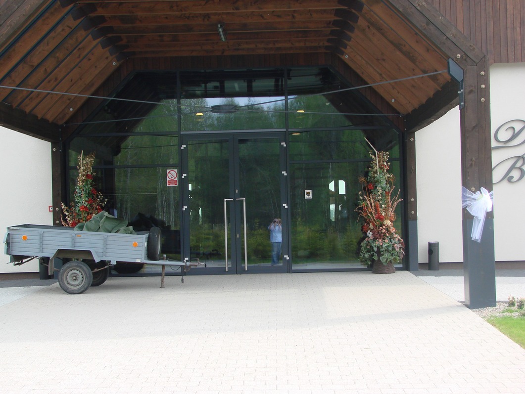 Duże szklane drzwi wejściowe do budynku w Sypniewie k. Więcborka, z widocznym odbiciem osoby wewnątrz. Po lewej stronie stoi przyczepka z ładunkiem przykrytym zielonym materiałem, a po prawej...