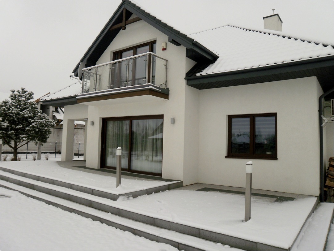 Nowoczesny dom jednorodzinny z białymi ścianami, balkonem z przezroczystą balustradą i dużymi przesuwnymi drzwiami tarasowymi, pokryty śniegiem.