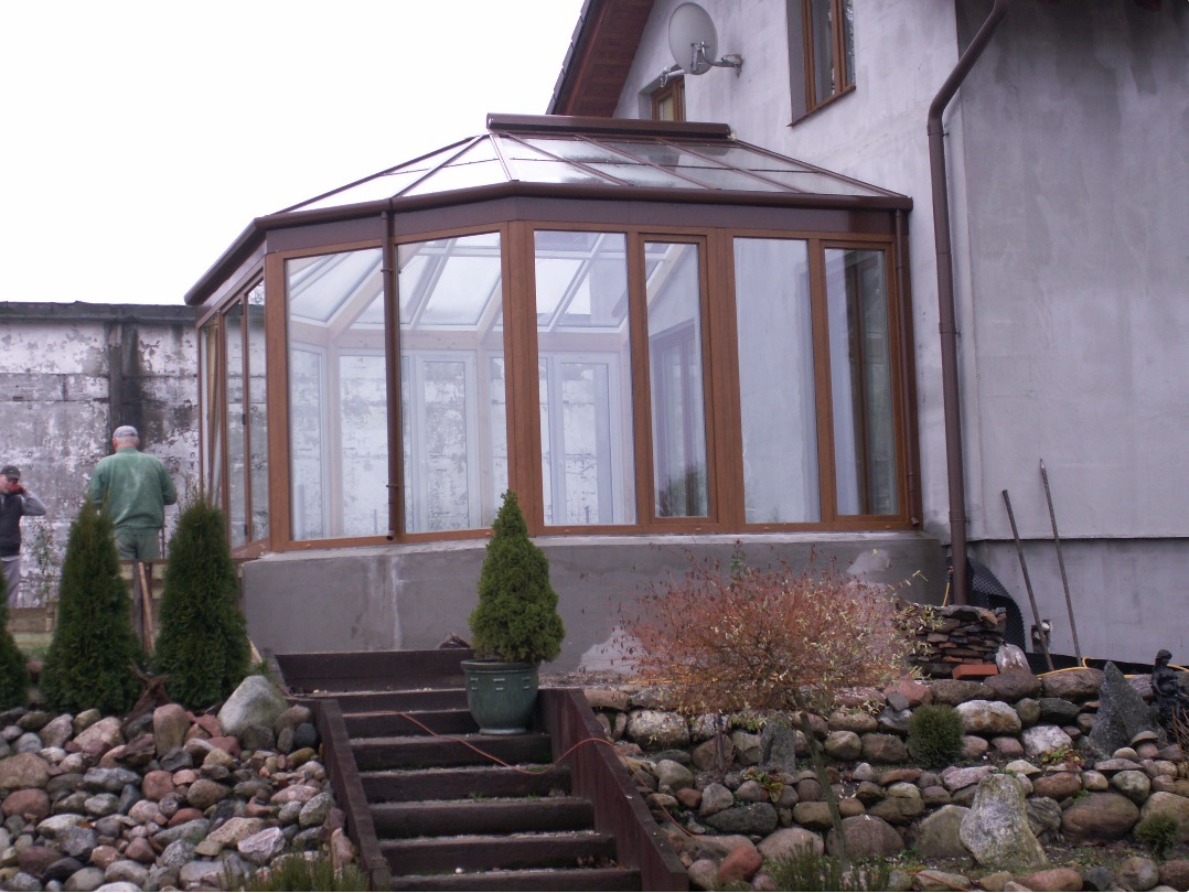Zewnętrzny widok przeszklonej oranżerii z brązowymi ramami, dobudowanej do szarego domu z widocznymi schodami wejściowymi i kamiennym ogrodem w tle.