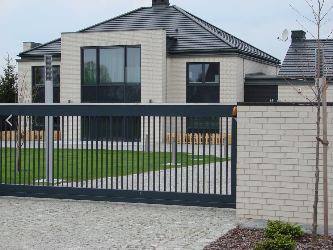 Nowoczesna, przesuwna brama w kolorze antracytowym przed domem z jasnej cegły, widok z poziomu chodnika wyłożonego kostką brukową.