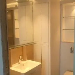 Nowoczesna łazienka z białą umywalką na szafce, lustrem z oświetleniem LED, zabudową meblową do sufitu i wiszącą toaletą w tle.