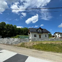 Nowoczesny dom z czarnym dachem i balkonem, otoczony kamiennym murem oporowym z szarego kamienia, widok z poziomu chodnika wyłożonego kostką.