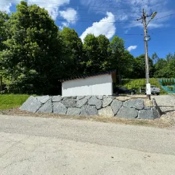 Nowo wybudowany murek oporowy z nieregularnych, szarych kamieni, zabezpieczający teren przy asfaltowej drodze, z białym budynkiem w tle i zielenią drzew na wzgórzu.