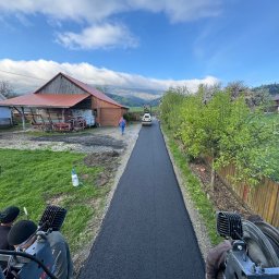 Firma Handlowo-Usługowa Marek Hajdas - Świeżo położony asfalt na wiejskiej drodze, widok z maszyny budowlanej, w tle budynki gospodarcze i osoba idąca w oddali.