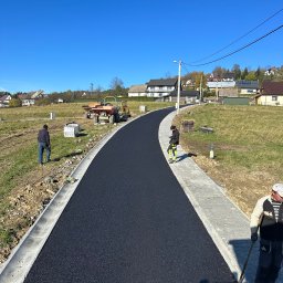 Firma Handlowo-Usługowa Marek Hajdas - Nowo wyasfaltowana droga w Lubieniu, z ułożoną kostką brukową po bokach, widoczni pracownicy i maszyny budowlane, błękitne niebo w tle.