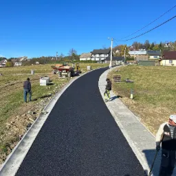Nowo wyasfaltowana droga w Lubieniu, z ułożoną kostką brukową po bokach, widoczni pracownicy i maszyny budowlane, błękitne niebo w tle.