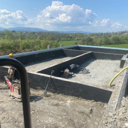 Firma Handlowo-Usługowa Marek Hajdas - Wykop fundamentowy z izolacją przeciwwilgociową ścian, widoczny ubijak wibracyjny, łopata i rury, w tle krajobraz wiejski z górami i błękitnym niebem.