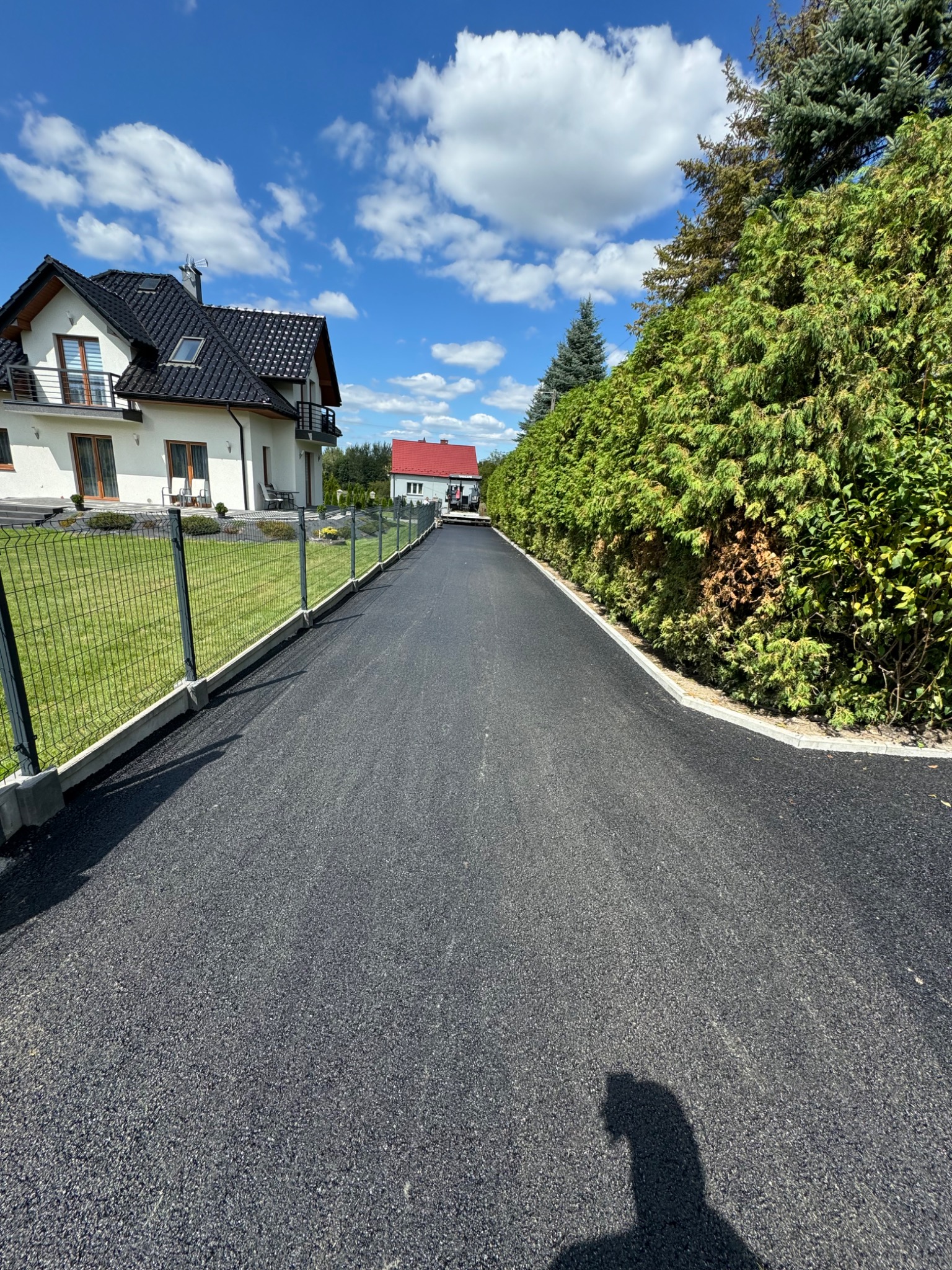 Nowa, czarna nawierzchnia asfaltowa prowadząca do domu z białymi ścianami i ciemnym dachem, z widocznym ogrodzeniem z siatki i zielonym żywopłotem po bokach, sfotografowana w słoneczny dzień.