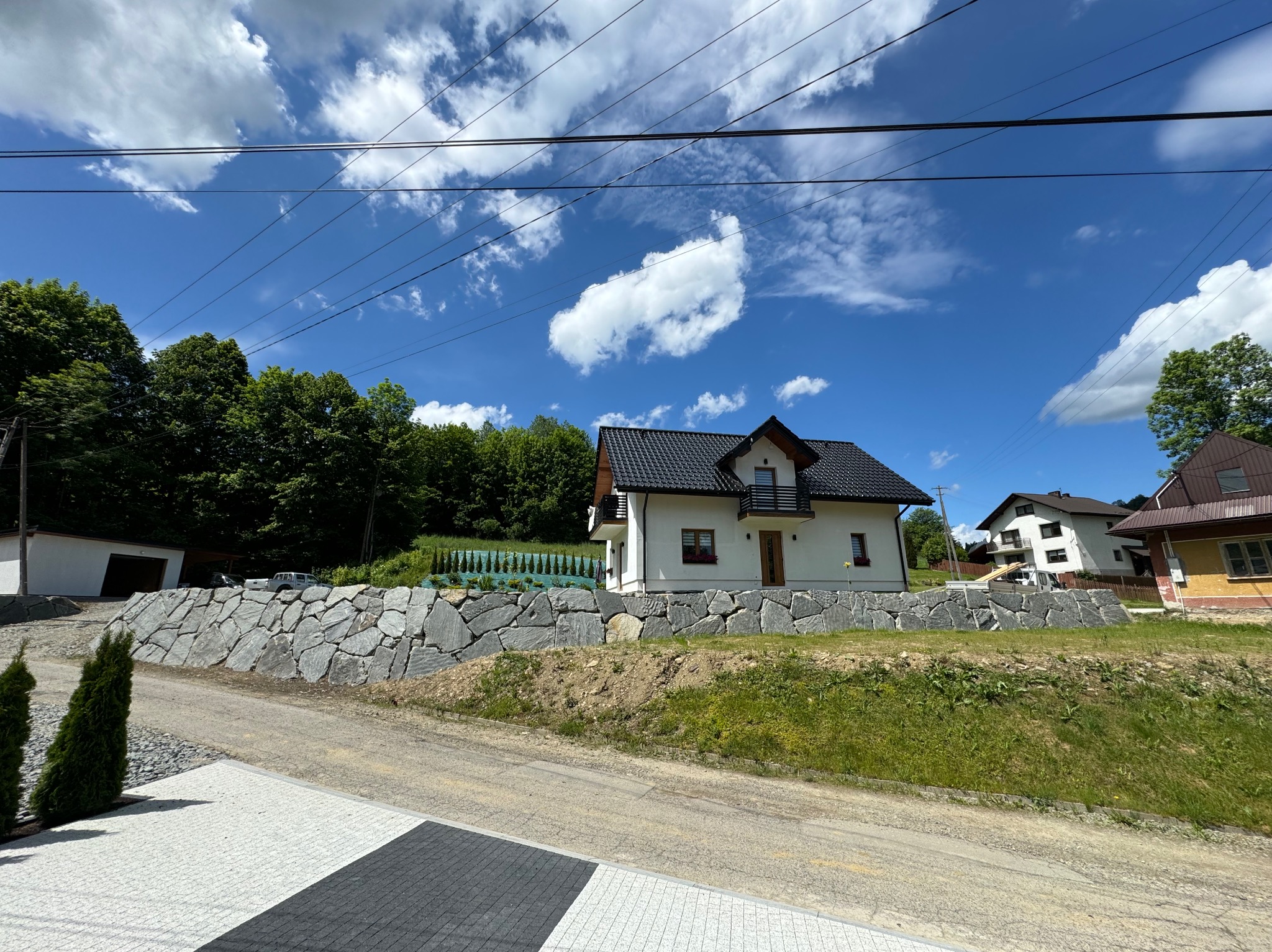 Nowoczesny dom z czarnym dachem i balkonem, otoczony kamiennym murem oporowym z szarego kamienia, widok z poziomu chodnika wyłożonego kostką.