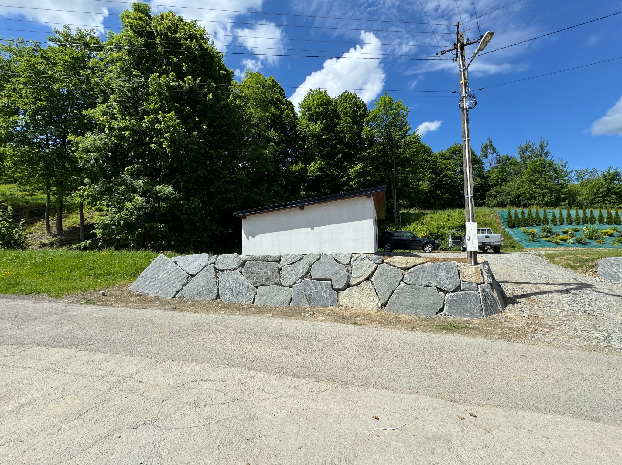 Nowo wybudowany murek oporowy z nieregularnych, szarych kamieni, zabezpieczający teren przy asfaltowej drodze, z białym budynkiem w tle i zielenią drzew na wzgórzu.