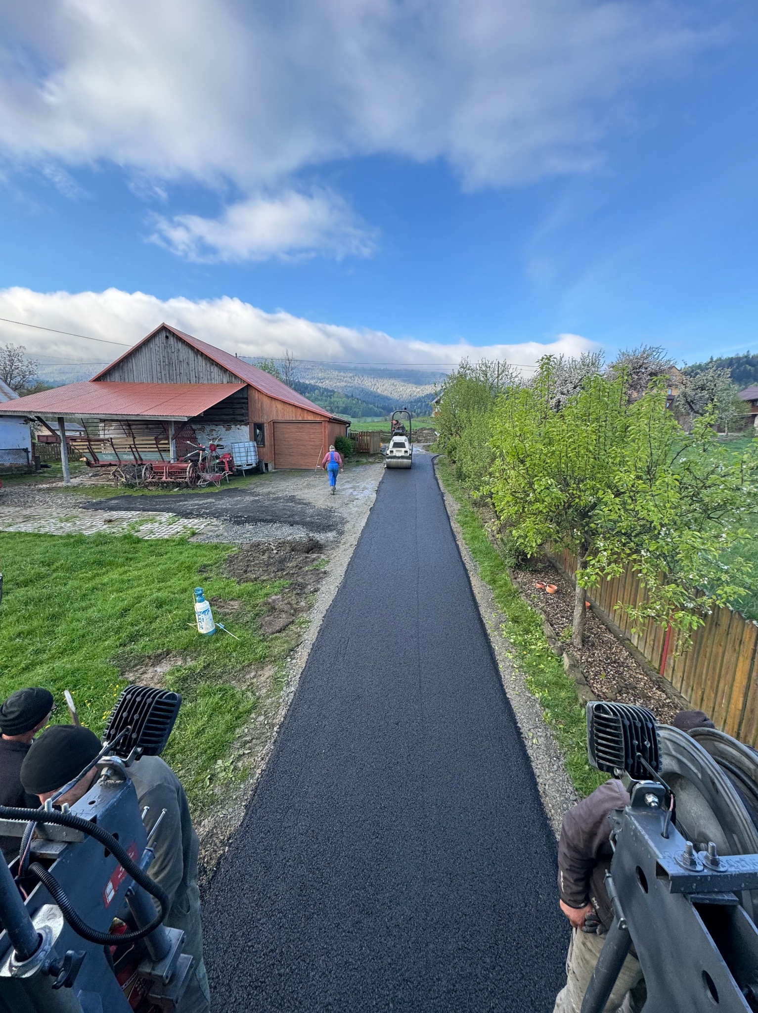 Świeżo położony asfalt na wiejskiej drodze, widok z maszyny budowlanej, w tle budynki gospodarcze i osoba idąca w oddali.