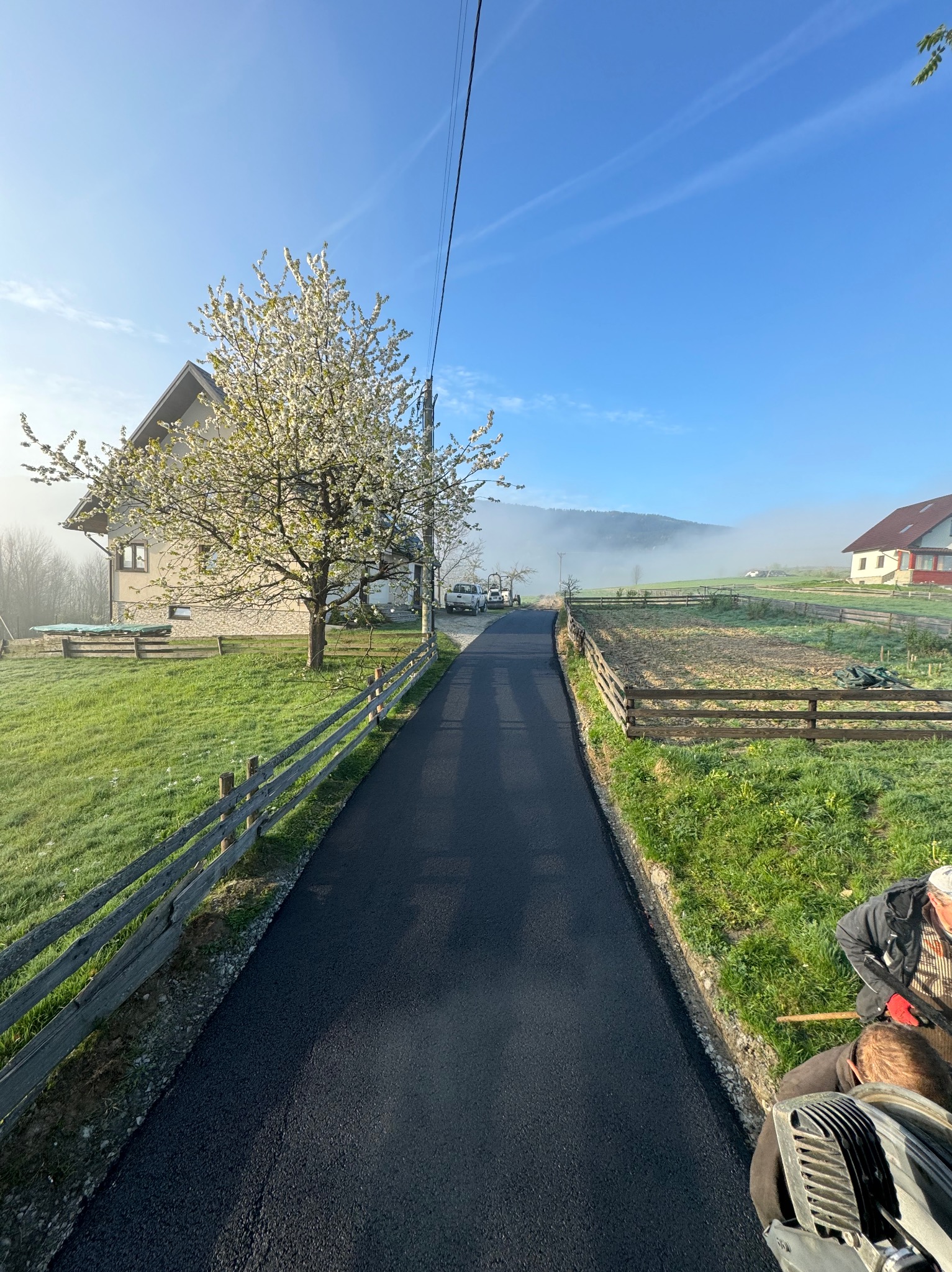 Nowo wyasfaltowana droga dojazdowa prowadząca do domu z kwitnącym drzewem, widoczna praca wykończeniowa obrzeży i lekka mgła w oddali.