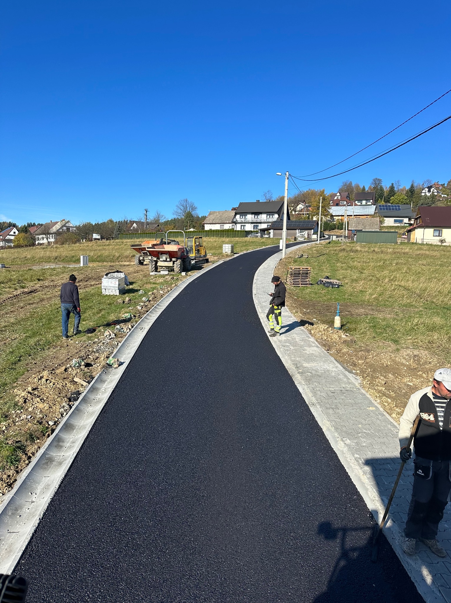 Nowo wyasfaltowana droga w Lubieniu, z ułożoną kostką brukową po bokach, widoczni pracownicy i maszyny budowlane, błękitne niebo w tle.