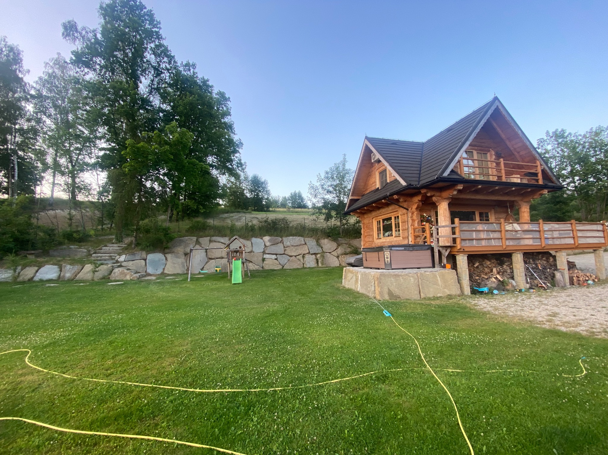 Drewniany dom z bali z tarasem i jacuzzi, otoczony zielonym trawnikiem i skalniakiem, widoczny plac zabaw dla dzieci.