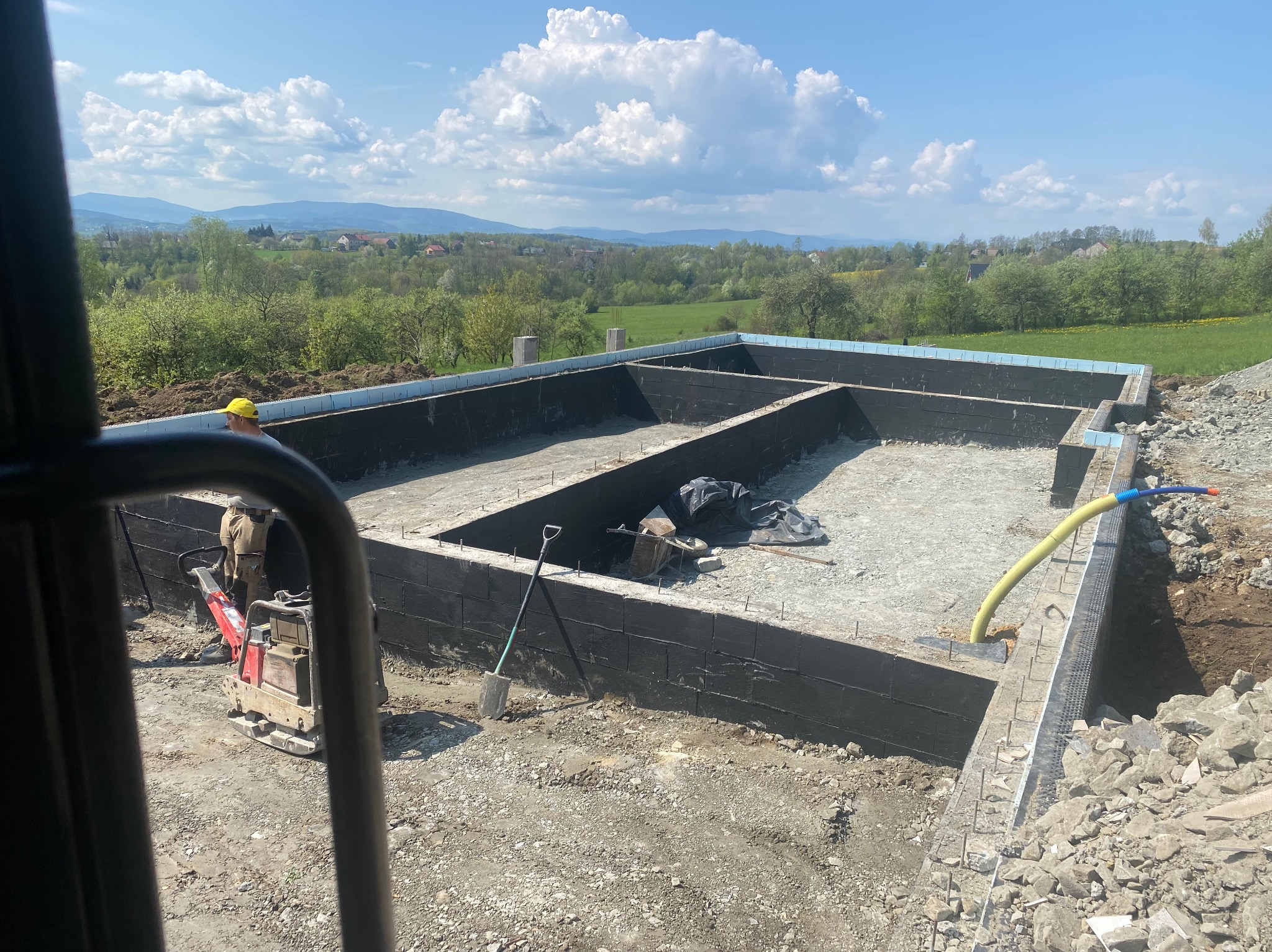 Wykop fundamentowy z izolacją przeciwwilgociową ścian, widoczny ubijak wibracyjny, łopata i rury, w tle krajobraz wiejski z górami i błękitnym niebem.
