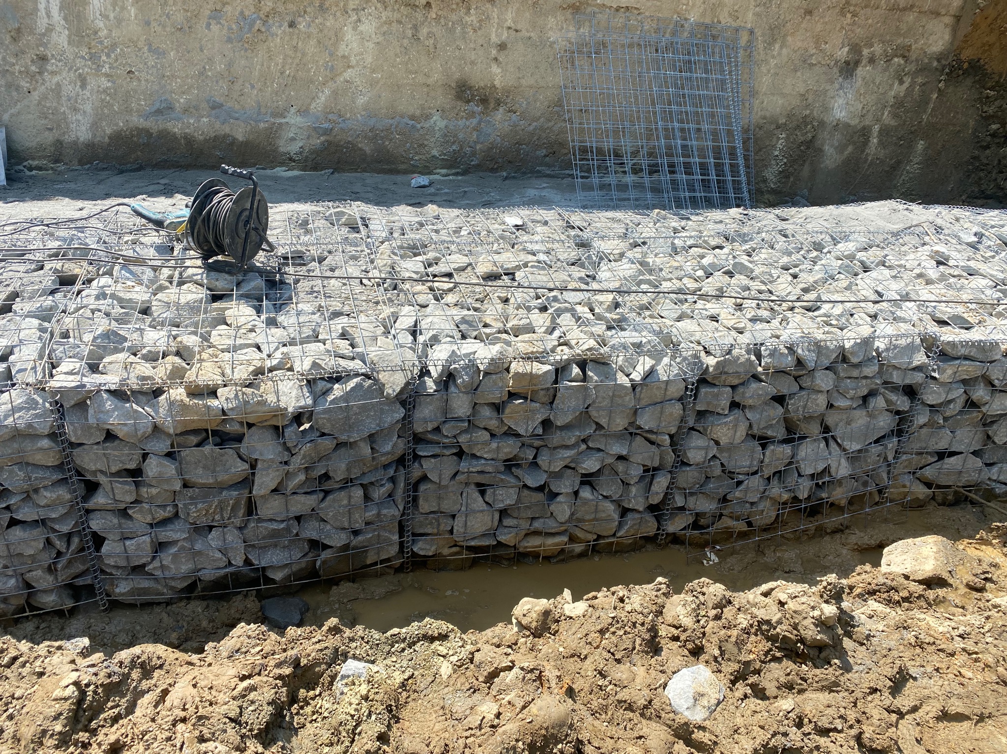 Konstrukcja z kamieni w metalowej siatce, prawdopodobnie gabion, w trakcie budowy, z widoczną ziemią i narzędziami w tle.