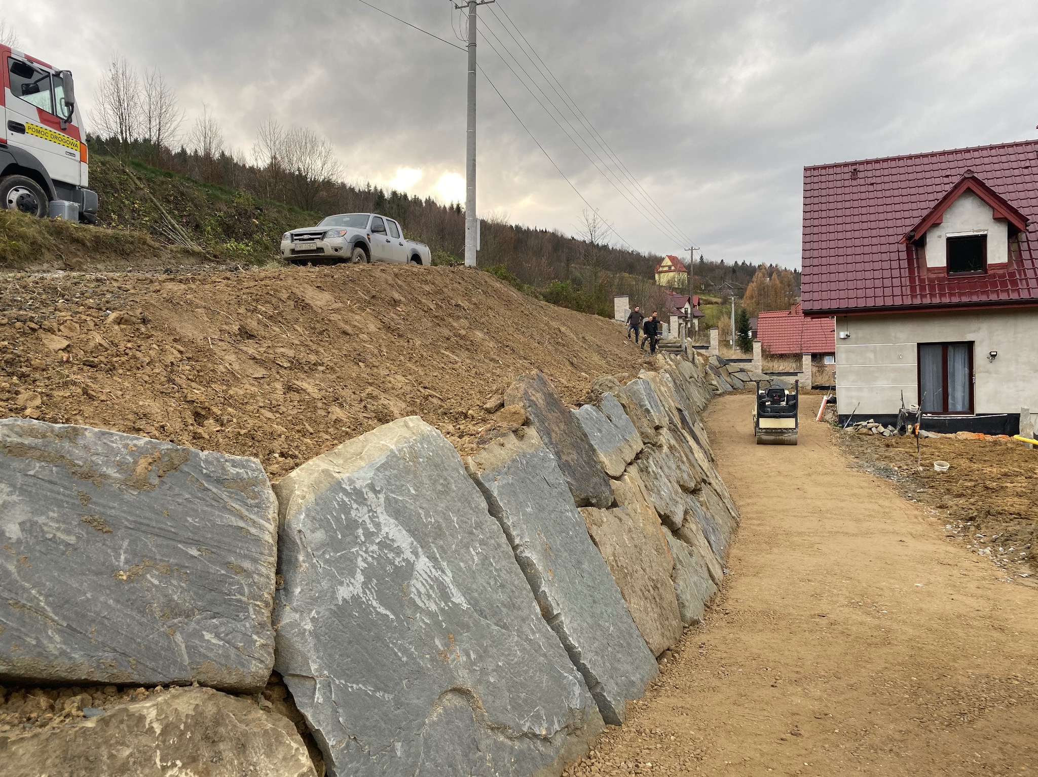 Wznoszenie kamiennego muru oporowego wzdłuż drogi gruntowej, ubijarka w tle, na wzgórzu zaparkowany samochód dostawczy, pochmurne niebo.