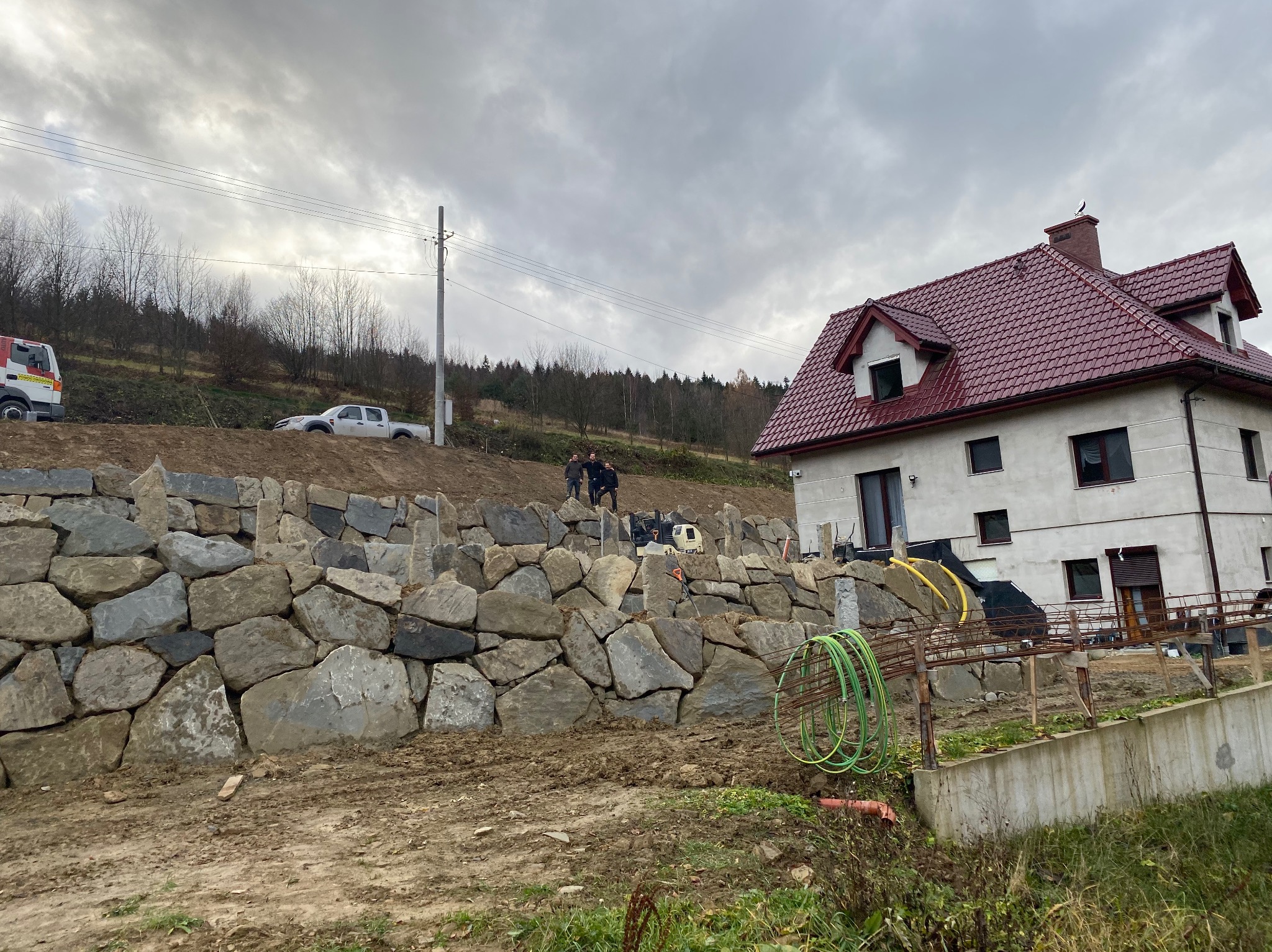 Wznoszenie kamiennego muru oporowego na nierównym terenie, widoczni pracownicy oraz budynek z czerwonym dachem w tle, pochmurne niebo.