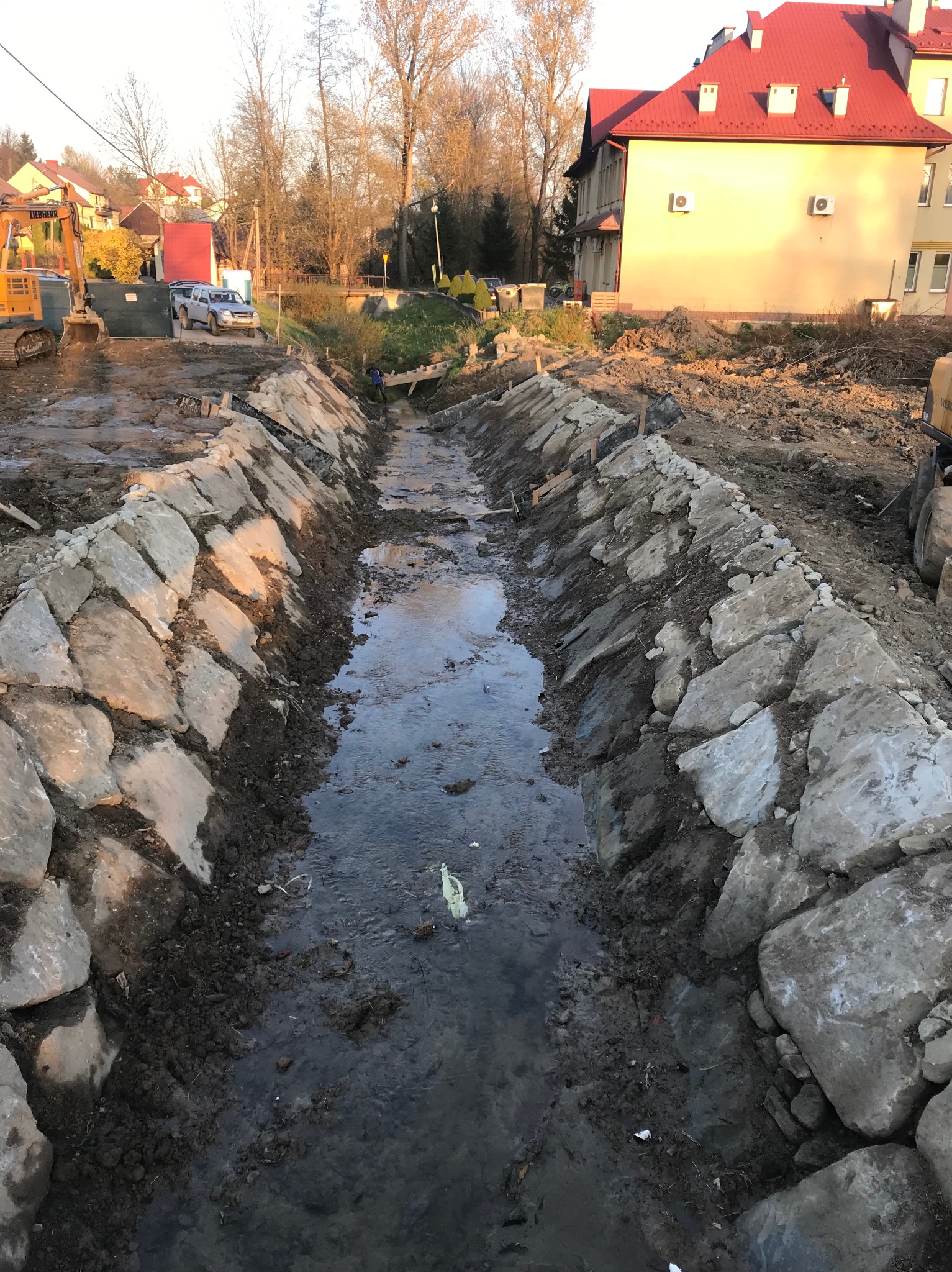 Budowa rowu melioracyjnego z kamiennym umocnieniem brzegów, w tle budynek mieszkalny i maszyny budowlane.