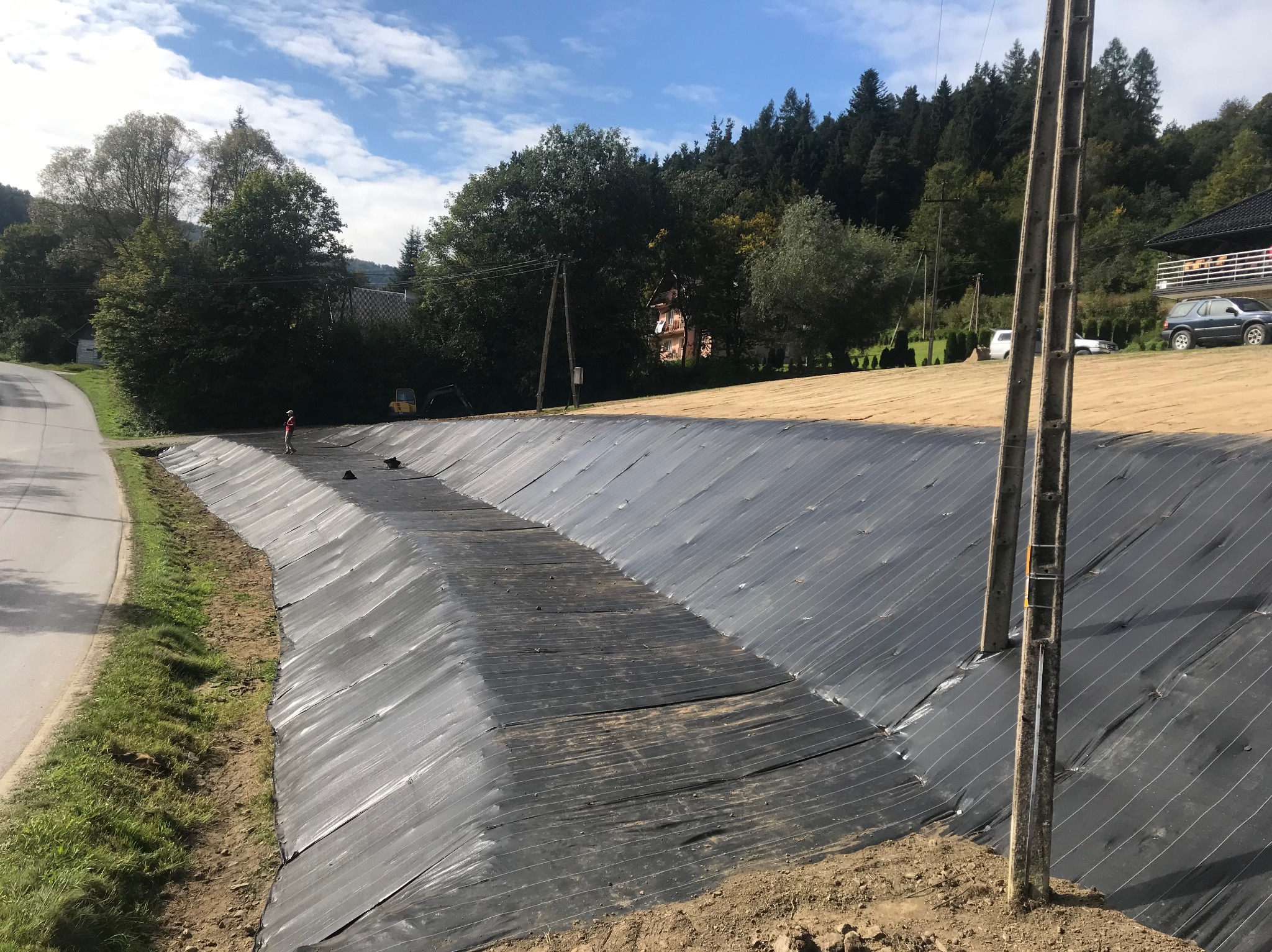 Skarpa zabezpieczona czarną geowłókniną, widoczna droga asfaltowa, trawa, drzewa, budynek mieszkalny i słupy energetyczne w tle, mała koparka i pracownik w oddali.