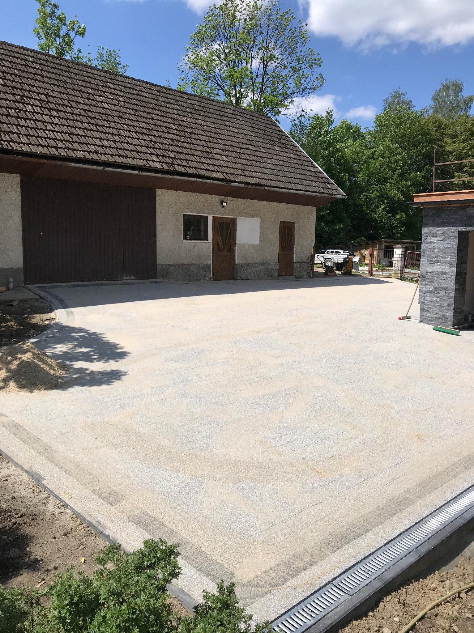 Nowo utwardzony, jasny żwirowy podjazd przed budynkiem gospodarczym z brązową bramą i dwojgiem drzwi, widoczny system odprowadzania wody wzdłuż krawędzi, częściowo zarośnięty trawą, zarys cienia...