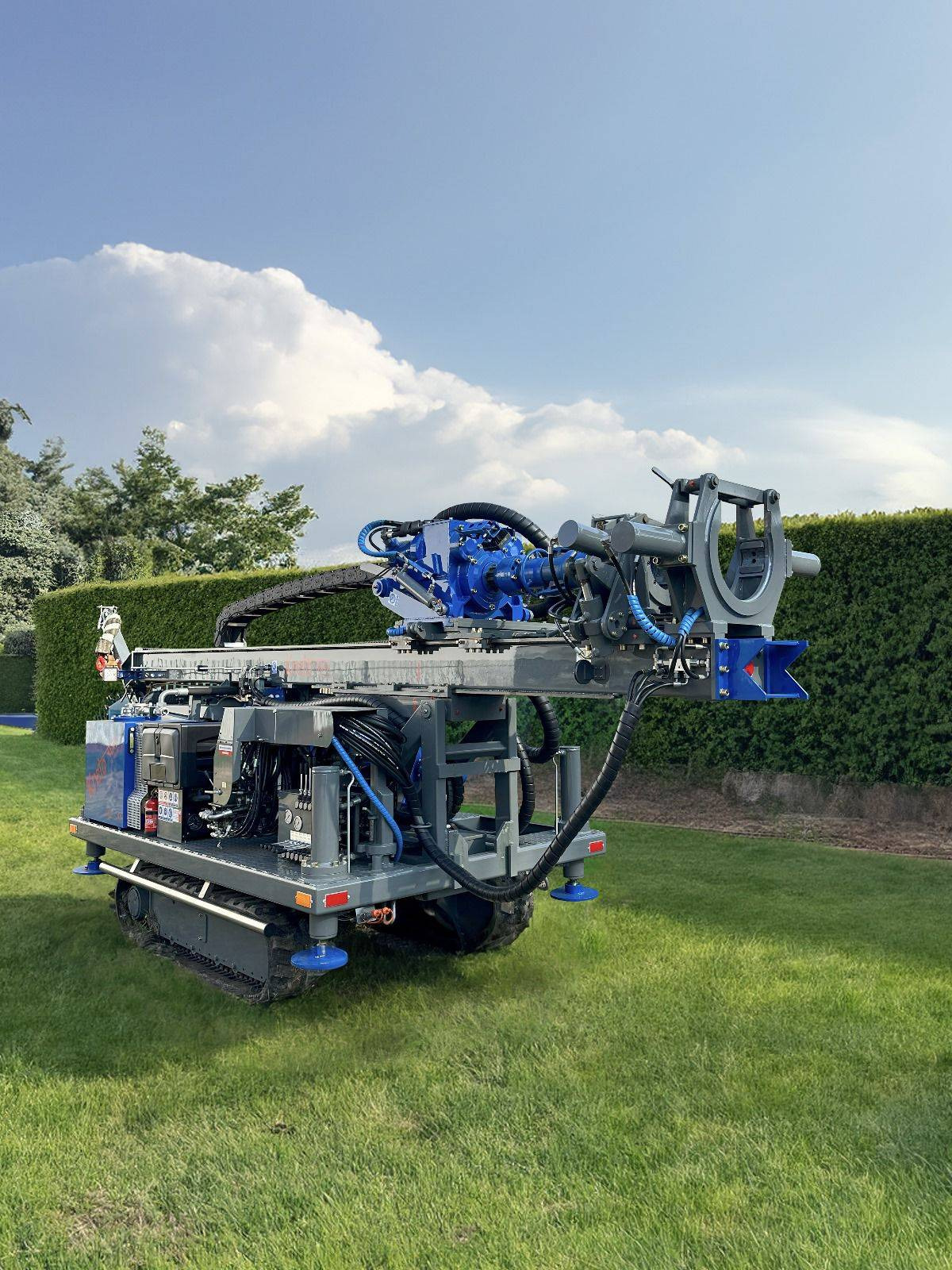 Mobilna wiertnica do studni głębinowych na trawiastym terenie, z niebieskimi akcentami i zielonym żywopłotem w tle, gotowa do pracy.