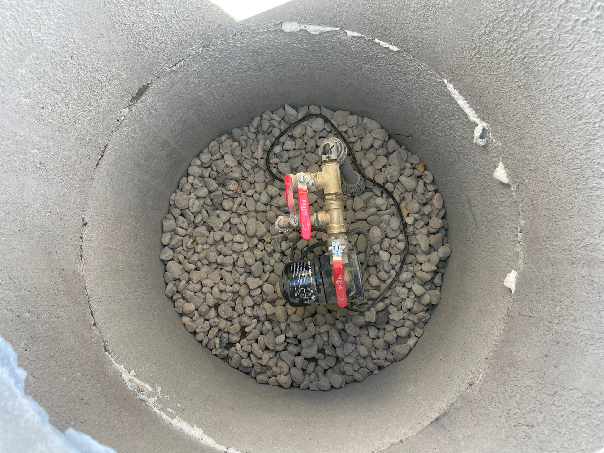 Wnętrze betonowej studni z pompą i zaworami na warstwie otoczaków. Widoczne uszczelnienia na ścianach studni. Ujęcie z góry.