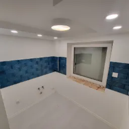 Wykończona łazienka z niebieską glazurą ułożoną do połowy ściany, widoczne przyłącza wodne i okno z surowym wykończeniem wnęki.