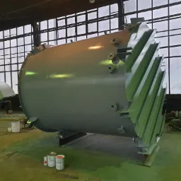 Pomalowany na szaro zbiornik przemysłowy z elementami montażowymi, stojący w hali produkcyjnej z dużymi oknami, obok widoczne puszki z farbą.