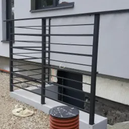 Czarna, metalowa konstrukcja zabezpieczająca taras przy budynku z elewacją w odcieniach szarości, widoczna z poziomu gruntu pokrytego żwirem.