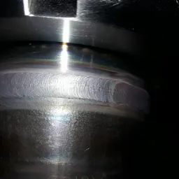 Szlifowana spoiną na okrągłym elemencie metalowym, widoczne tęczowe zabarwienia na powierzchni stali nierdzewnej.