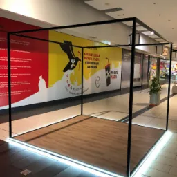 Metalowa konstrukcja w centrum handlowym, oświetlona diodami LED, na podłodze imitacja drewna, w tle reklama społeczna dotycząca problemu plastiku w oceanach.