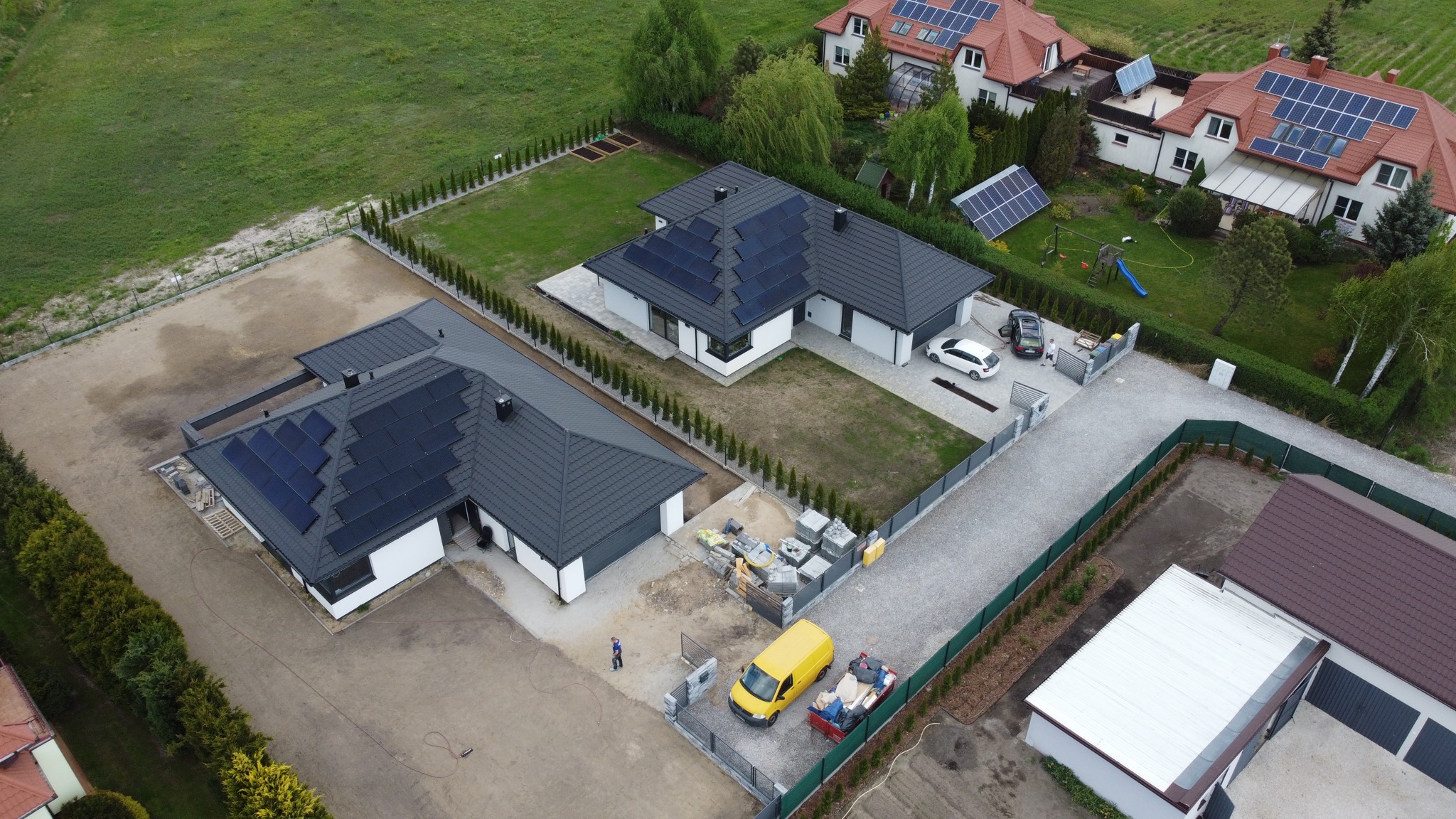 Widok z drona na dwa nowoczesne domy z panelami słonecznymi na dachach, z żółtym vanem i materiałami budowlanymi na posesji jednego z nich.