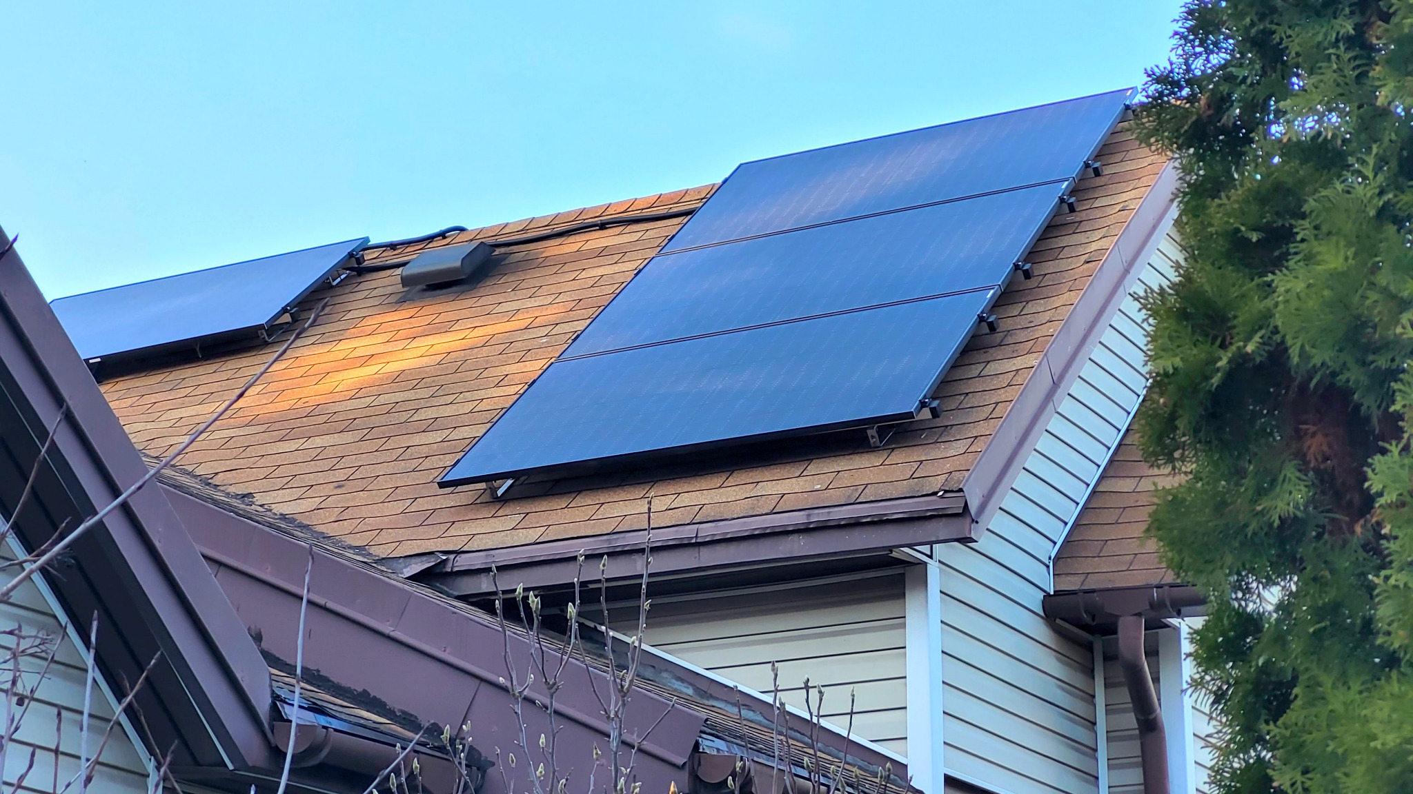 Panele słoneczne na brązowym dachu domu jednorodzinnego, widoczne elementy montażowe i część instalacji, obok iglaste drzewo.