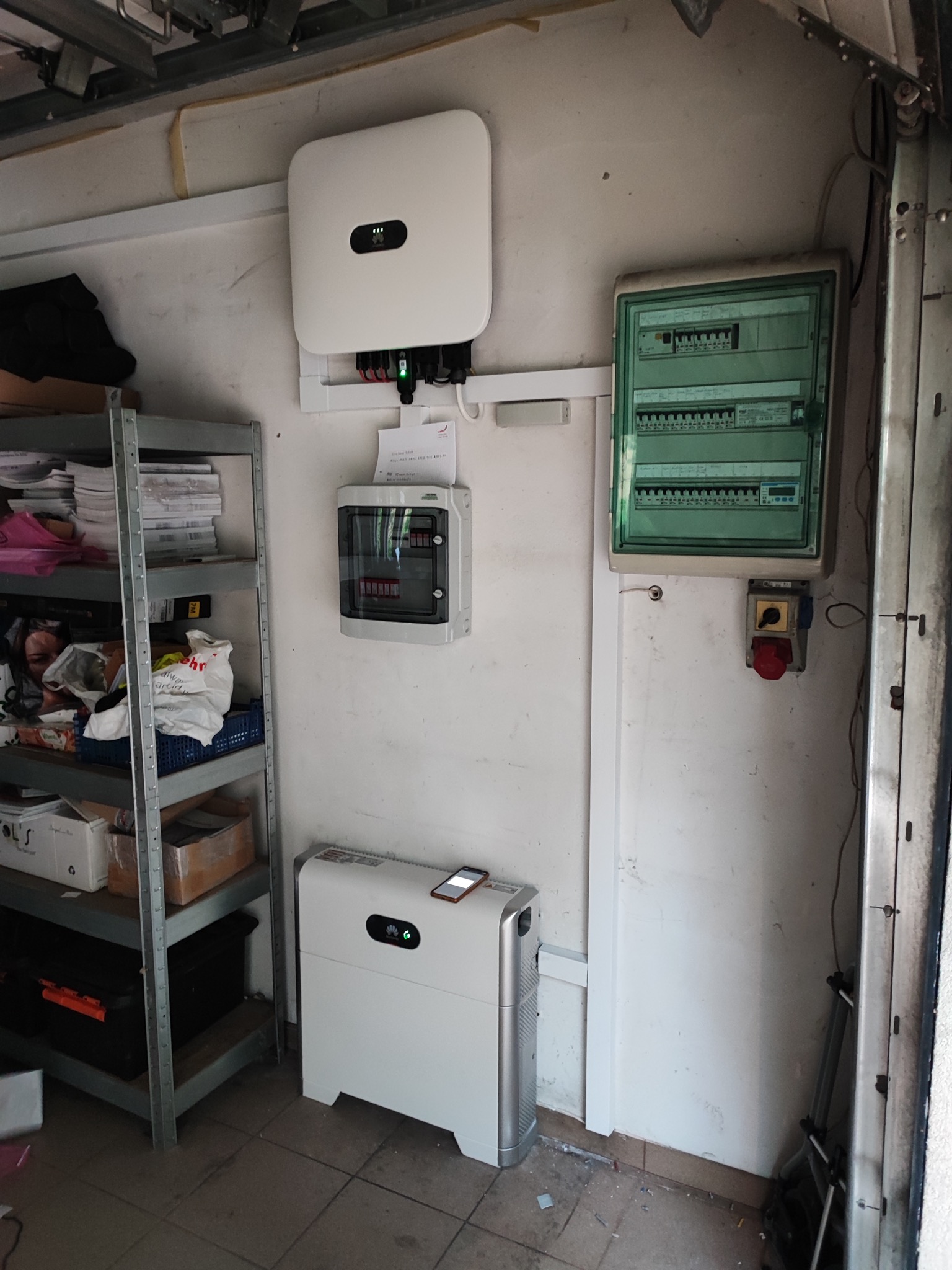 Biały inwerter fotowoltaiczny Huawei zamontowany na ścianie garażu, obok rozdzielnica elektryczna w zielonej obudowie i półka z różnymi przedmiotami. Na inwerterze leży telefon.