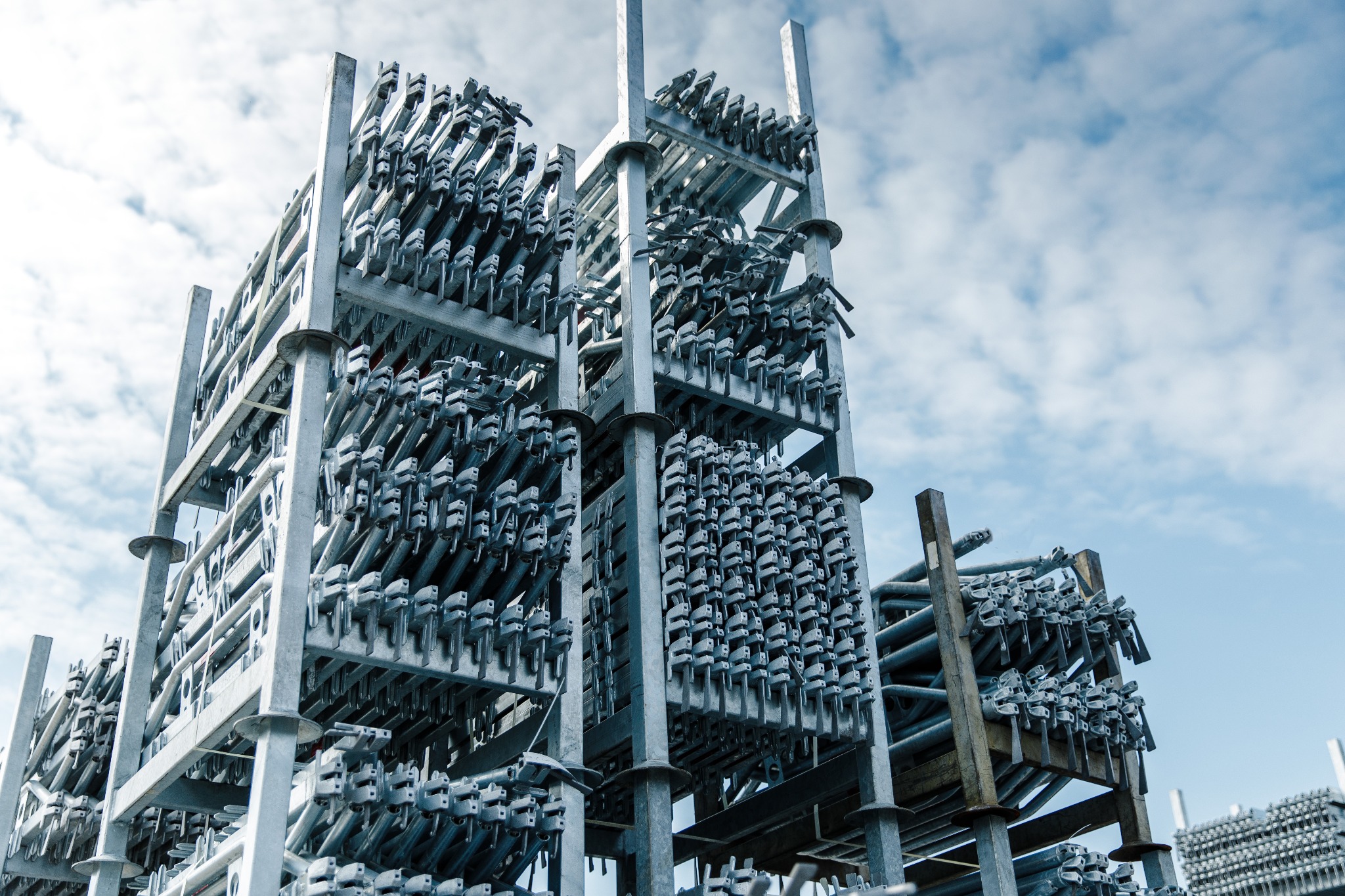 Metalowe rusztowania modułowe, składowane w Ostrzeszowie, widoczne na tle błękitnego nieba z chmurami.