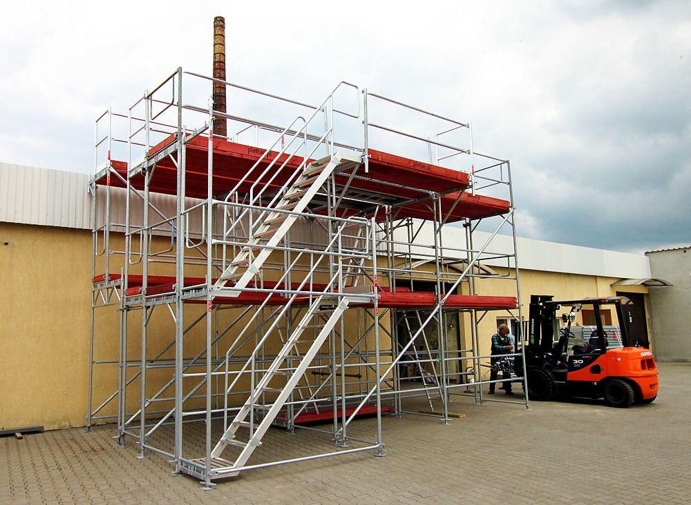 Wielopoziomowe rusztowanie z czerwonymi podestami i schodami, obok stoi pomarańczowy wózek widłowy z operatorem.