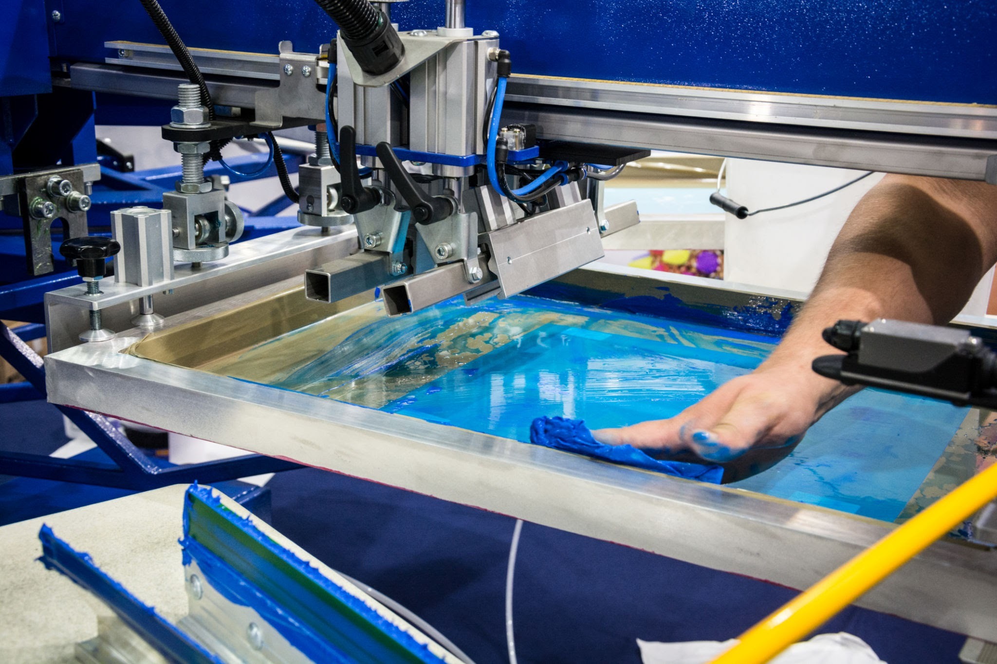 Czyszczenie ramy sitodrukarskiej z resztek farby za pomocą niebieskiej szmatki, widoczny fragment koszulki i elementy maszyny do sitodruku.