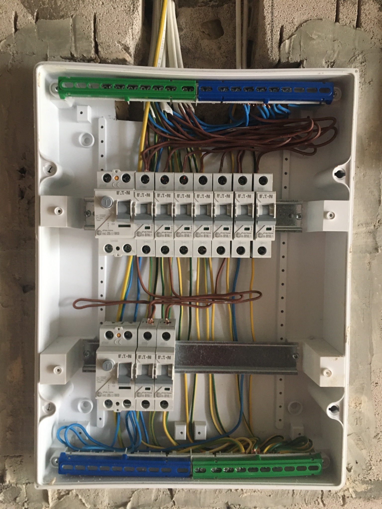 Wnętrze rozdzielnicy elektrycznej z zamontowanymi bezpiecznikami EATON, widoczne kolorowe przewody i listwy zaciskowe, tło stanowi surowa ściana z betonu komórkowego.