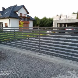 Nowoczesna, pozioma brama wjazdowa z szarych, metalowych profili na tle domu z ciemną dachówką i budującego się garażu.