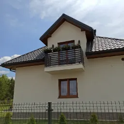 Dom jednorodzinny z balkonem ozdobionym kwiatami, widoczny fragment ogrodzenia panelowego z siatki.