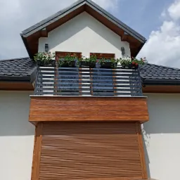 Nowoczesny balkon z poziomymi, metalowymi barierkami, ozdobiony kwiatami w donicach, z brązową roletą zewnętrzną na dole.