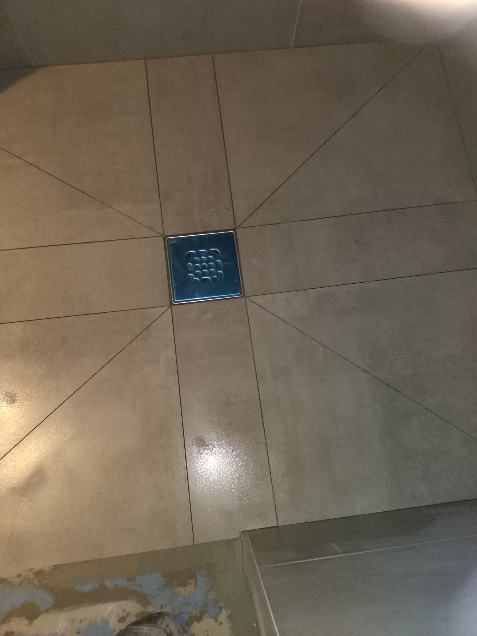 Beżowe płytki podłogowe ułożone promieniście wokół kwadratowego odpływu liniowego z niebieską metalową kratką, widoczne fragmenty podłoża z resztkami kleju.