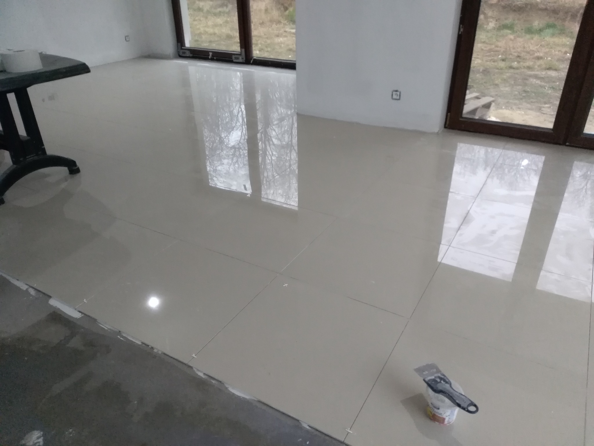 Jasne, błyszczące płytki podłogowe ułożone w pomieszczeniu z dużymi oknami, widoczne narzędzia glazurnicze i krzyżyki dystansowe.