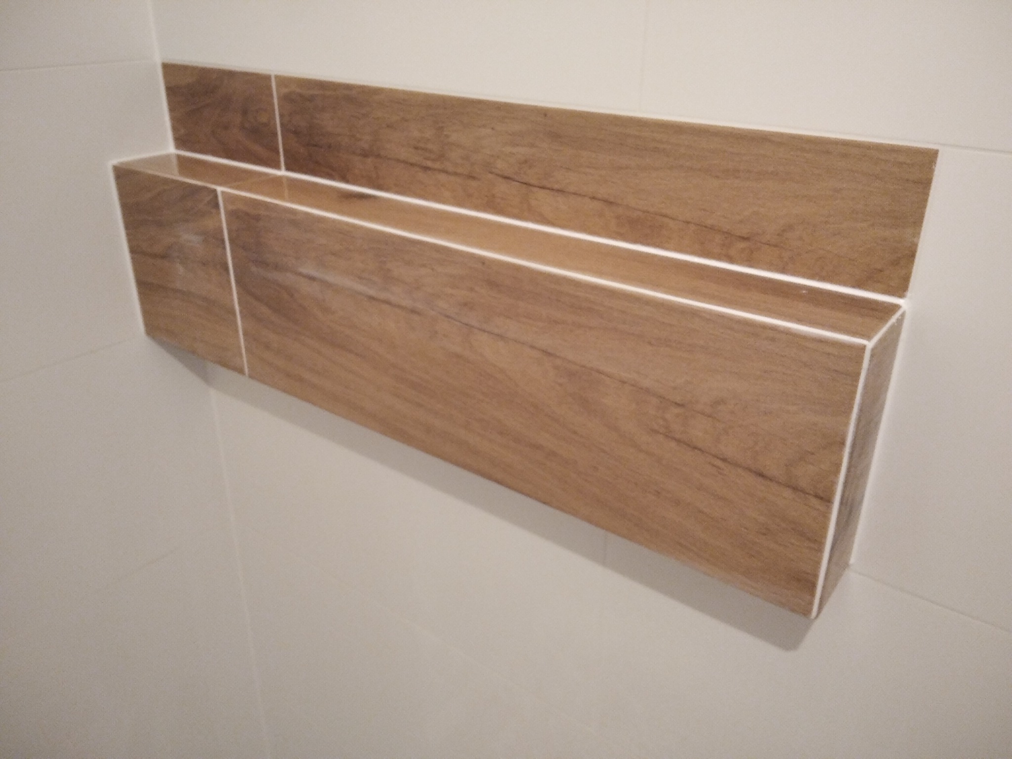 Półka ścienna w łazience, wykonana z płytek imitujących drewno, z białymi fugami, zamontowana na białych płytkach ściennych.