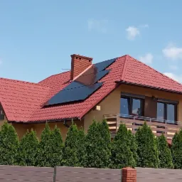 Dom jednorodzinny z czerwonym dachem i trzema panelami słonecznymi zamontowanymi na połaci dachu, widok z poziomu ogrodzenia z tujami.