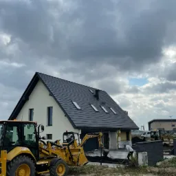 Żółta koparka CAT 428 stoi na nierównym terenie budowy przed domem jednorodzinnym z ciemną dachówką, w tle pochmurne niebo i inne maszyny budowlane.