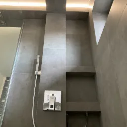 Nowoczesna kabina prysznicowa z szarymi płytkami, wbudowanymi półkami i oświetleniem LED, widoczna bateria Grohe.